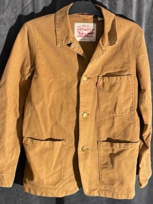Beige jacka från Levi's - Säljer en beige jacka från Levi's med klassisk krage, tre stora utanpåfickor och guldfärgade knappar. Jackan är tillverkad i slitstark bomull och har en enkel, rak passform som funkar till många olika stilar. I fint begagnat skick. 