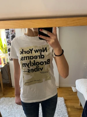Vit t-shirt med tryck från Stradivarius - Säljer en vit t-shirt från Stradivarius i storlek XS. T-shirten har ett stort tryck framtill med texten 'New York dreams. Brooklyn vibes' i svart och guld. Klassisk passform och rund halsringning. Perfekt till jeans eller shorts.