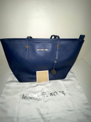 Blå axelväska från Michael Kors - Snygg marinblå axelväska från Michael Kors i skinn med guldfärgade detaljer och dragkedja upptill. Väskan har justerbara axelband och ett klassiskt MK-hänge framtill. Perfekt storlek för allt du behöver ha med dig och stilren design som passar till det mesta.