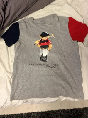 Grå Polo Bear t-shirt från Ralph Lauren - Säljer en grå t-shirt från Polo Ralph Lauren med ikoniskt Polo Bear-tryck på bröstet. T-shirten har korta ärmar i olika färger, en marinblå och en röd, vilket ger en unik look. Tillverkad i mjuk bomull och har normal passform.