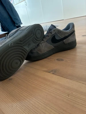 Grå Nike Air Force 1 mockasneakers - Säljer ett par grå Nike Air Force 1 sneakers i mocka och textil. Skorna har mörkgrå sula, svart swoosh och grå snörning. Låg modell med klassisk rund tå och diskreta blå detaljer på häl och plös. Perfekta för dig som gillar stilrena och tidlösa sneakers. 