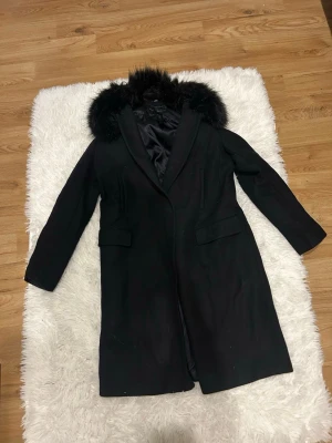 Svart kappa med fuskpäls från Zara - Säljer en svart lång kappa från Zara Basic med snygg fuskpäls runt kragen. Klassisk rak modell med två fickor fram och diskreta knappar. Perfekt för dig som vill ha en stilren och varm jacka till höst och vinter.