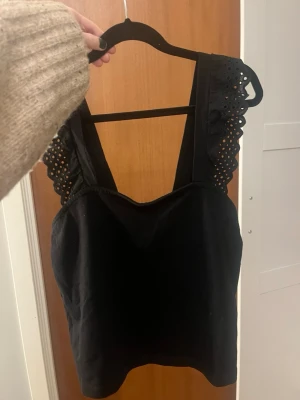 Svart linnetopp med spets från H&M - Säljer min jättefina svarta topp då den tyvärr aldrig kommer till användning. Toppen har breda axelband med spetsdetaljer och hålmönster. Den är croppad och har en rak, enkel passform som är lätt att styla till jeans eller kjol. Endast använd en gång💕