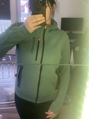 Hoddie husqvarna - Säljer en grön hoddie från husqvarna med hel dragkedja och huva. Tröjan har svarta detaljer, orange snörning och dragkedjor på fickorna samt en bröstficka. 