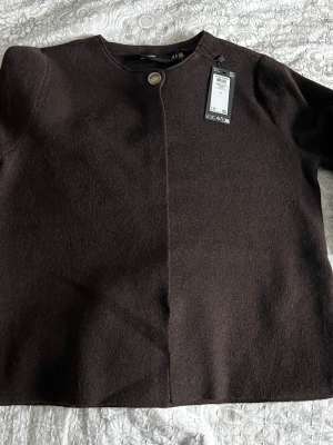 Oanvänd Brun kofta från Vero Moda, M - Mörkbrun kofta från Vero Moda i storlek M. Enkel design med rund halsringning och en stor knapp framtill. Tillverkad i mjukt material som passar perfekt till lager på lager under höst och vinter.