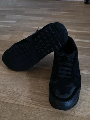 Valentino Rockrunner svarta sneakers - Säljer ett par svarta Valentino Rockrunner sneakers med coola nitar på hälen och diskret kamouflagemönster på ovandelen. Skorna har snörning, rund tå och är tillverkade i en mix av mocka, skinn och textil. Perfekta för dig som gillar en edgy och stilren look.
