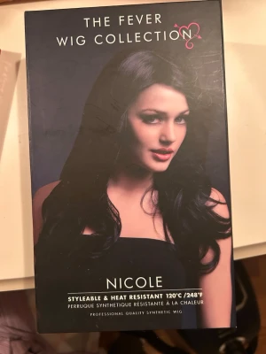 Svart syntetisk peruk Nicole 66 cm - Säljer en svart syntetisk peruk från Fever Wig Collection, modell Nicole. Peruken är 66 cm lång med mjuka vågor och sidbena. Den är värmetålig upp till 120°C och har justerbar passform samt naturlig look med fejkhud vid benan.