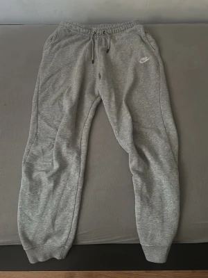 Grå mjukisbyxor från Nike med snörning - Säljer ett par grå mjukisbyxor från Nike i storlek S. Byxorna har resår i midjan med snörning, muddar vid bensluten och två sidofickor. Klassisk Nike-logga broderad på vänster ben. Perfekta för chill dagar eller träning.