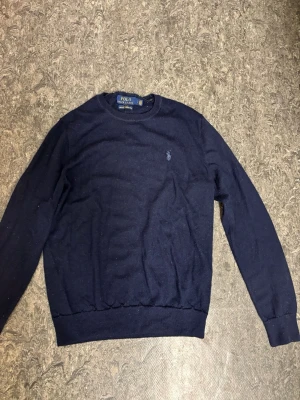 Mörkblå merinoulltröja Polo Ralph Lauren - Klassisk mörkblå tröja från Polo Ralph Lauren i mjuk merinoull. Tröjan har rund halsringning, långa ärmar och den ikoniska broderade loggan på bröstet. Perfekt för dig som gillar stilrena och tidlösa plagg.