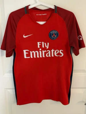 Röd PSG fotbollströja Nike - Snygg röd Paris Saint-Germain fotbollströja från Nike med Fly Emirates-tryck på bröstet och klubbmärke. Tröjan har mörkröda ärmar med diskreta ränder och blå detaljer vid kragen och sidorna. Perfekt för dig som älskar fotboll och PSG.