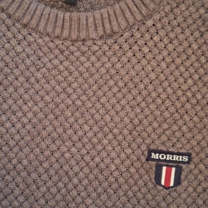 Grå stickad tröja från Morris - Säljer en stickad tröja från Morris med grov struktur och rund hals. Tröjan har ett broderat Morris-märke på bröstet och ribbade muddar vid ärmslut och nederkant. Perfekt för dig som gillar klassisk och stilren look. Har funnits ett hål vid ena axeln som är professionellt lagat av skräddare. Syns inte om man inte vet om det. Se bilder.
