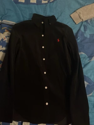 Svart skjorta med röd Polo Ralph Lauren märke - Säljer en svart  skjorta med röd ralph lauren märke från Polo Ralph Lauren, helt ny använd med klassisk krage och vita knappar. Skjortan har lång ärm och den ikoniska röda loggan broderad på bröstet. Perfekt för dig som gillar stilrena och griscg plagg.