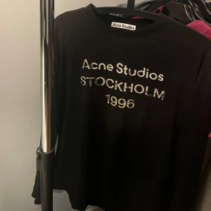 Svart långärmad tröja Acne Studios - Svart långärmad tröja från Acne Studios med vit text 'Acne Studios Stockholm 1996' framtill. Klassisk rund halsringning och avslappnad passform. Perfekt för dig som gillar stilrena och trendiga plagg med tydlig logga.