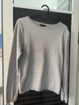 Ljusgrå stickad tröja från LMTD - En enkel och stilren ljusgrå stickad tröja från LMTD. Tröjan har rund halsringning, långa ärmar och ribbade muddar vid hals, ärmslut och nederkant. Perfekt till lager på lager under höst och vinter.