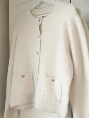 stickad kofta  - Mysig vit kofta med fluffig textur och rund hals. Koftan har dekorativa knappar framtill och två fickor med knappdetaljer. Perfekt för lager på lager under kyligare dagar.