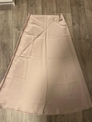 Beige långkjol i satin - Säljer en stilren beige långkjol i mjukt och glansigt satintyg. Kjolen har en enkel A-linjeformad siluett och faller snyggt. Perfekt för dig som gillar minimalistisk och elegant stil.