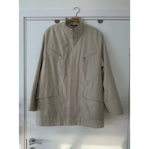 Beige parkajacka med dragkedjor - Snygg beige parkajacka med hög krage och flera dragkedjeförsedda fickor. Jackan har långa ärmar och diskret mönstrad yta. Perfekt för dig som gillar stilren och funktionell design.