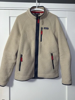  Patagonia fleece - Säljer en beige fleecejacka från Patagonia i storlek M. Jackan har hel dragkedja, tre fickor med dragkedja och röda detaljer. Mjuk och varm med hög krage och mörkblå kantband. Perfekt för kyliga dagar och outdoor vibes.