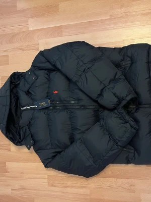 Svart pufferjacka från Polo Ralph Lauren - Säljer en svart pufferjacka från Polo Ralph Lauren med huva och dragkedja. Jackan har det klassiska röda Polo-broderiet på bröstet och flera praktiska fickor med dragkedja. Perfekt för kalla dagar och riktigt snygg till streetwear-stilen.
