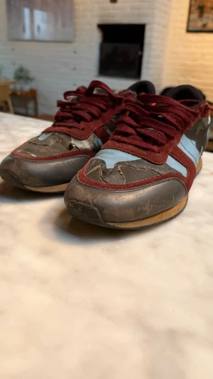 Valentino Rockrunner sneakers camo - Säljer ett par Valentino Rockrunner sneakers med camomönster i blått, svart, grönt och brunt. Skorna har vinröda mockadetaljer, röda skinnhällor bak och svarta nitar på hälen. Snygga snörningar och platt sula. Materialmix av mocka, skinn och textil. Väldigt schyssta till hösten. De har lite slitningar men inget som syns bra och de går alltid såklart att fixa tilldet