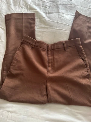 Bruna kostymbyxor från Gina Tricot - Snygga bruna kostymbyxor från Gina Tricot i storlek 38. Byxorna har raka ben, bälteshällor och sidofickor fram samt en dekorativ bakficka. Tillverkade i ett mjukt bomullsliknande material med klassisk passform. Lite noppriga