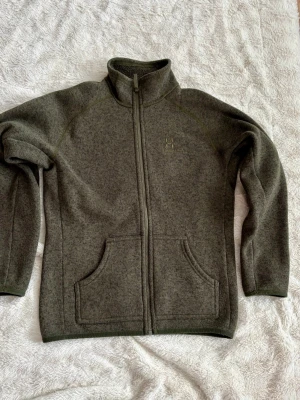 Mörkgrön fleecekofta från Haglöfs - Mysig mörkgrön fleecekofta från Haglöfs med hel dragkedja och två stora fickor framtill. Hög krage för extra värme och enkel, stilren design. Perfekt för kyliga dagar och outdoor-häng.