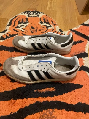 Adidas Samba sneakers vit/svart - Klassiska Adidas Samba sneakers i vitt skinn med svarta ränder och grå mockadetaljer på tån. Skorna har den ikoniska blå Adidas-loggan på plösen och gummisula. Perfekta för dig som gillar retrostil och streetwear. Obs ingen sula och inga snörren 