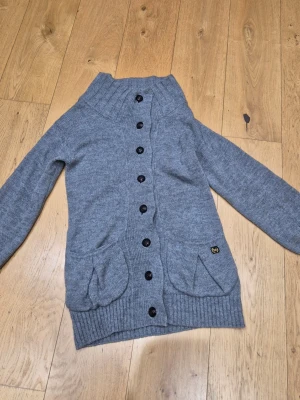 Grå stickad kofta från Burberry st S - Mysig grå stickad kofta från Thomas Burberry med stora svarta knappar hela vägen, två fickor fram och ribbad krage. Riktigt varm med merinoull och alpackaull 