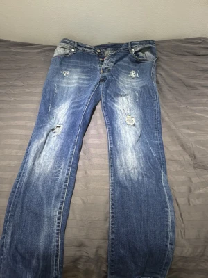 Dsquared2 blå slitna jeans - Säljer ett par blå jeans från Dsquared2 med coolt sliten look och distinkta slitningar framtill och baktill. Jeansen har en klassisk femficksmodell, rak passform och grönt Dsquared2-märke bak i midjan. Materialet är robust denim i bomull.