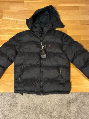 Svart pufferjacka från Polo Ralph Lauren - Säljer en svart pufferjacka från Polo Ralph Lauren med huva och dragkedja framtill. Jackan har två sidofickor med dragkedja och den klassiska röda loggan på bröstet. Perfekt för kalla dagar och går att matcha till typ allt. Hör av dig om du har nån fundering😊👍