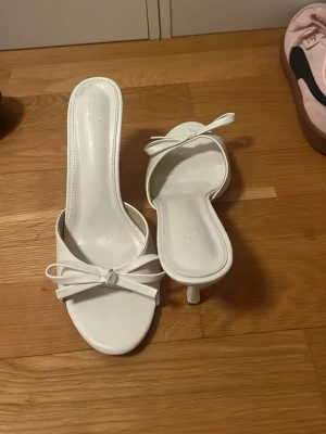 Vita sandaler med rosett och klack - Säljer ett par vita sandaler från Satin med öppen tå, smal hög klack och en söt rosettdetalj framtill. Skorna har en elegant och minimalistisk design i syntetmaterial, perfekta för att lyfta vilken outfit som helst.