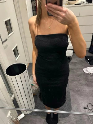 Svart glittrig axelbandslös klänning - Säljer en svart, glittrig axelbandslös klänning i tight modell. Klänningen är midilång och har en snygg slits bak för extra rörelsefrihet. Perfekt för dig som vill sticka ut med en elegant och stilren look.