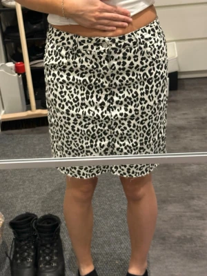 Leopardmönstrad kortkjol med knappar - Säljer en snygg kortkjol med leopardmönster i svart och vitt. Kjolen har knappar framtill och klassiska fickor. Perfekt för dig som vill sticka ut lite extra och gillar djurmönster. Passar till många olika stilar och är enkel att matcha.