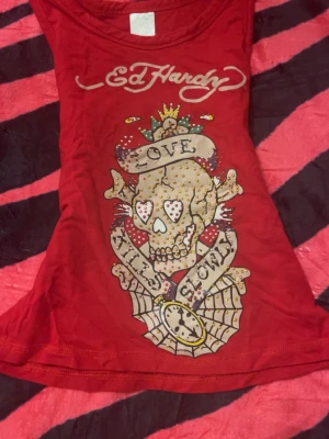 Röd Ed Hardy linnetopp med rhinestones - Säljer en röd linnetopp från Ed Hardy (som tyvärr inte passade mig) med  ikoniskt dödskalletryck, texten 'Love Kills Slowly' och massor av färgglada strasstenar. Toppen har ärmlös design och logga både fram och bak. Perfekt för dig som gillar statement-plagg och edgy stil.