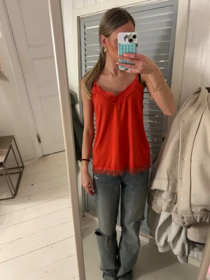 Röd linnetopp med spets från Freequent - Säljer en röd linnetopp från Freequent i storlek XS. Toppen har tunna axelband och fina spetsdetaljer längs både ringning och nederkant. Perfekt att styla med jeans eller kjol för en trendig look.