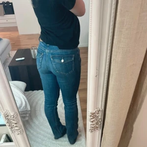 Blå bootcut jeans med fickdetaljer - Säljer ett par klassiska blå lågmidjade jeans med bootcut passform. Jeansen har snygga fickor bak med knappdetaljer och sitter riktigt bra i midjan. Fint skick☺️