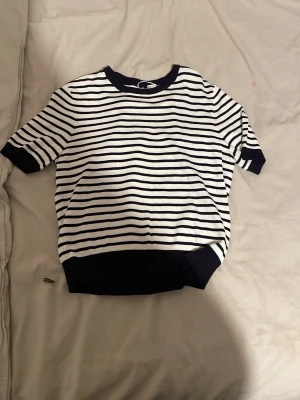 Randig kortärmad topp i bomull - Säljer en vit och marinblå randig topp med korta ärmar och ribbade muddar. Toppen har en klassisk rund halsringning och är gjord i mjuk bomull, perfekt för en avslappnad stil.