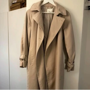 Beige kappa med bälte - trenchcoat liknande - Snygg beige trenchcoat med klassisk krage, knytskärp i midjan och detaljerade ärmslut med spännen. Lång modell som ger en clean och tidlös look. Perfekt för lager-på-lager och funkar till alla outfits.