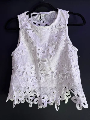 Vit blommig spetslinne från NLY Trend - Superfin vit ärmlös topp från NLY Trend i storlek 36. Toppen har broderade blommor i spets och en snygg, lite vågig kant nertill. Perfekt för sommarens alla tillfällen och ger en fräsch och romantisk look.