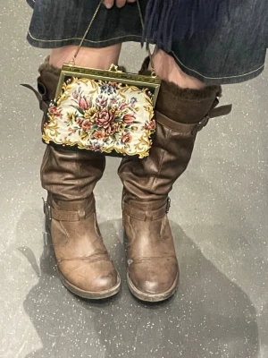 Blommig vintage kuvertväska med knäppspänne - Säljer en unik vintage väska med broderat blommönster i flera färger och guldfärgad metallram. Väskan har en klassisk form, liten kedja som handtag och stängs med ett metallspänne.  Väskan är 19 cm bred och 15 cm hög. Den är i fint skick för vad den är men har lite repor på metallen likt de du ser på bild 7. Jag har använt den som en lite nicshad handväska/över axeln väska 😁