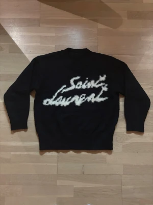 Svart Saint Laurent stickad tröja M - Säljer en svart stickad tröja från Saint Laurent i storlek M. Tröjan har ett stort vitt, fluffigt Saint Laurent-tryck på bröstet och ribbade muddar vid ärmslut och nederkant. Perfekt för dig som vill ha en statement piece med lyxig känsla.