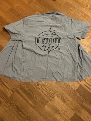 Ljusblå Carhartt skjorta med Detroit-tryck - Säljer en ljusblå skjorta från Carhartt med stort Detroit-tryck och blixtar på ryggen samt mindre Carhartt-logga på bröstet. Skjortan har korta ärmar, klassisk krage och knäppning framtill. Perfekt för dig som gillar streetwear och vill sticka ut.