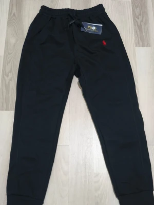 Svarta tracksuit byxor från Polo Ralph Lauren - Svarta tracksuit byxor från Polo Ralph Lauren med röd broderad logga på vänster sida. Byxorna har resår och snörning i midjan samt ribbade muddar vid bensluten. Tillverkade i mjukt bomullsmaterial för maximal komfort.