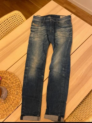 Blå Diesel jeans med slitningar - Säljer ett par blå jeans från Diesel med snygga slitningar och tvättade detaljer. Klassisk femficksmodell med Diesel Industry-märke på myntfickan. Jeansen har smal passform och raka ben, tillverkade i slitstarkt bomullsmaterial. Längd 100cm och bredd 40cm. Skriv vid frågor eller för fler bilder.