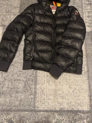 Svart pufferjacka Parajumpers Sheen S - Säljer en svart pufferjacka från Parajumpers, modell Sheen. Jackan har huva, dragkedja framtill, ribbade muddar och ikonisk gul detalj vid halsen. Fodrad insida och två fickor med dragkedja. Perfekt för kalla dagar och riktigt snygg streetstil. 100% äkta.