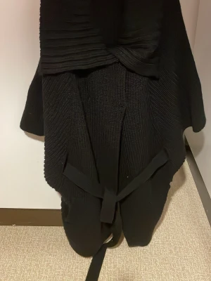 Svart stickad kofta med bälte - Säljer en svart stickad kofta med bälte i midjan. Koftan har en snygg struktur i tyget och är lite längre i modellen, perfekt att slänga på sig när det är kyligt. Den har en omlottkänsla och är riktigt mysig att bära. Ylle akryl o ull. Sk pingvinärm. Säljs på fler sidor.