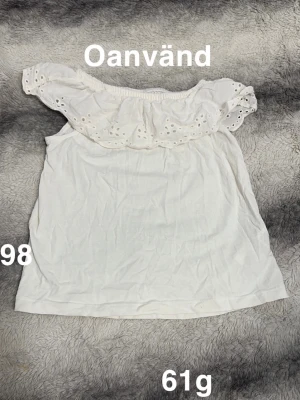 Vit offshoulder blus med volang - Säljer en söt vit blus i barnstorlek 98 med offshoulder-design och bred volang upptill. Volangen har broderade hålmönster som ger en romantisk känsla. Perfekt för sommarens alla tillfällen. Materialet känns mjukt och lätt.🌸 Oanvänd. Storlek 98