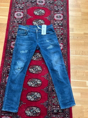 Dondup jeans  - Säljer ett par blå skinny jeans från Dondup med slitningar och färgstänk för en cool, avslappnad look. Jeansen har klassisk femficksdesign, låg midja och är tillverkade i mjukt denimtyg. Perfekta för dig som gillar streetstyle och detaljer.