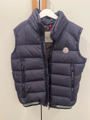 Mörkblå dunväst från Moncler - Snygg mörkblå dunväst från Moncler med hög krage och två dragkedjeförsedda fickor. Västen har quiltad design och Moncler-logga på bröstet. Perfekt för lager-på-lager och har sportiga ränder längst ner. Se sista bilden för en liten liten defekt som knappt syns 