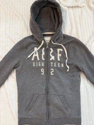 Hoodie zip up Abercrombie unik - Grå Abercrombie & Fitch-hoodie med dragkedja i vintagestil. Storlek M passar en vanlig S bättre! I toppskick! Har den vita Abercrombie & Fitch-logotypen i paljetter över bröstet.